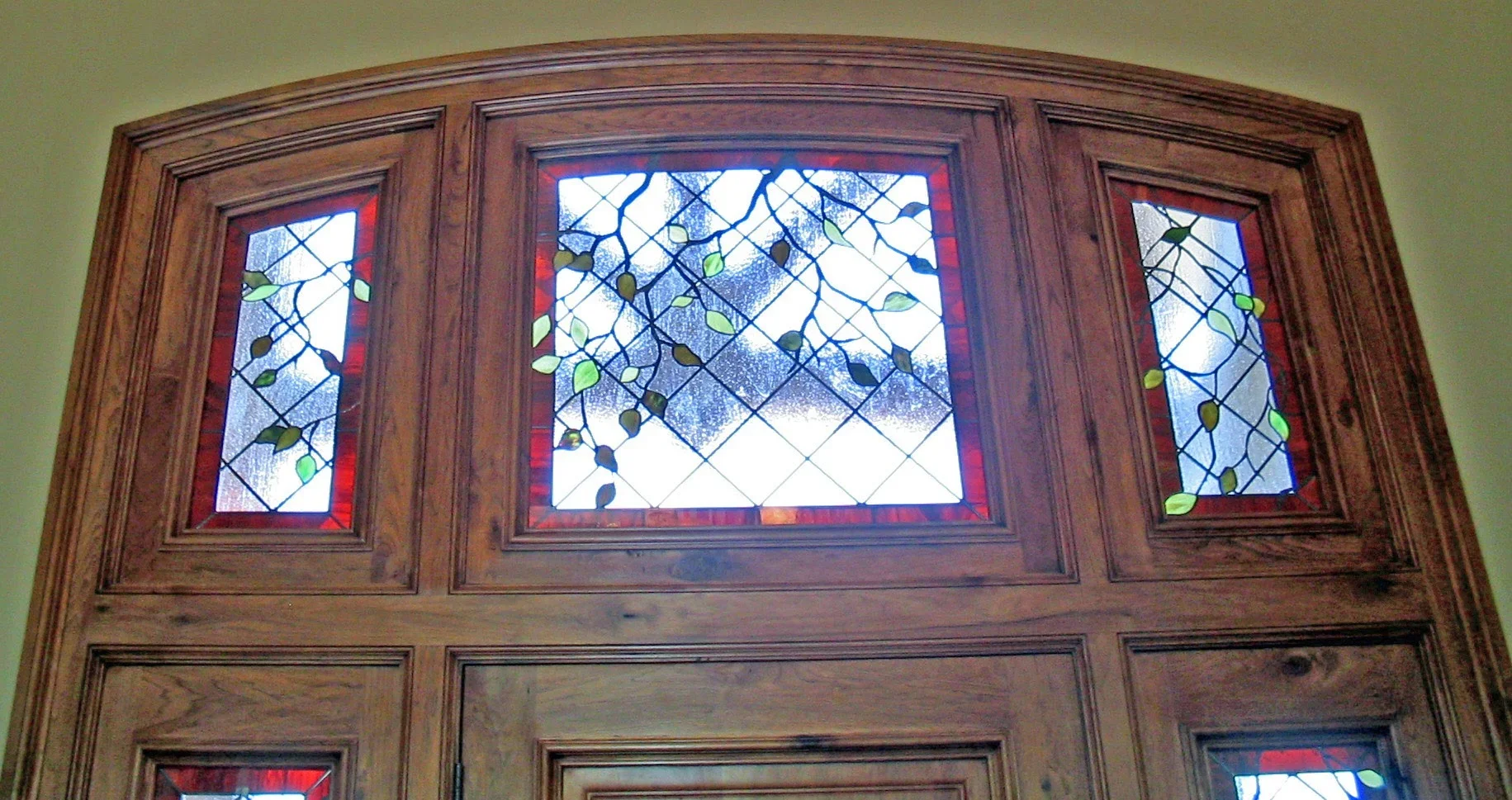 Arched stained glass transom with three panels featuring blue vines and green leaves on white glass with red borders