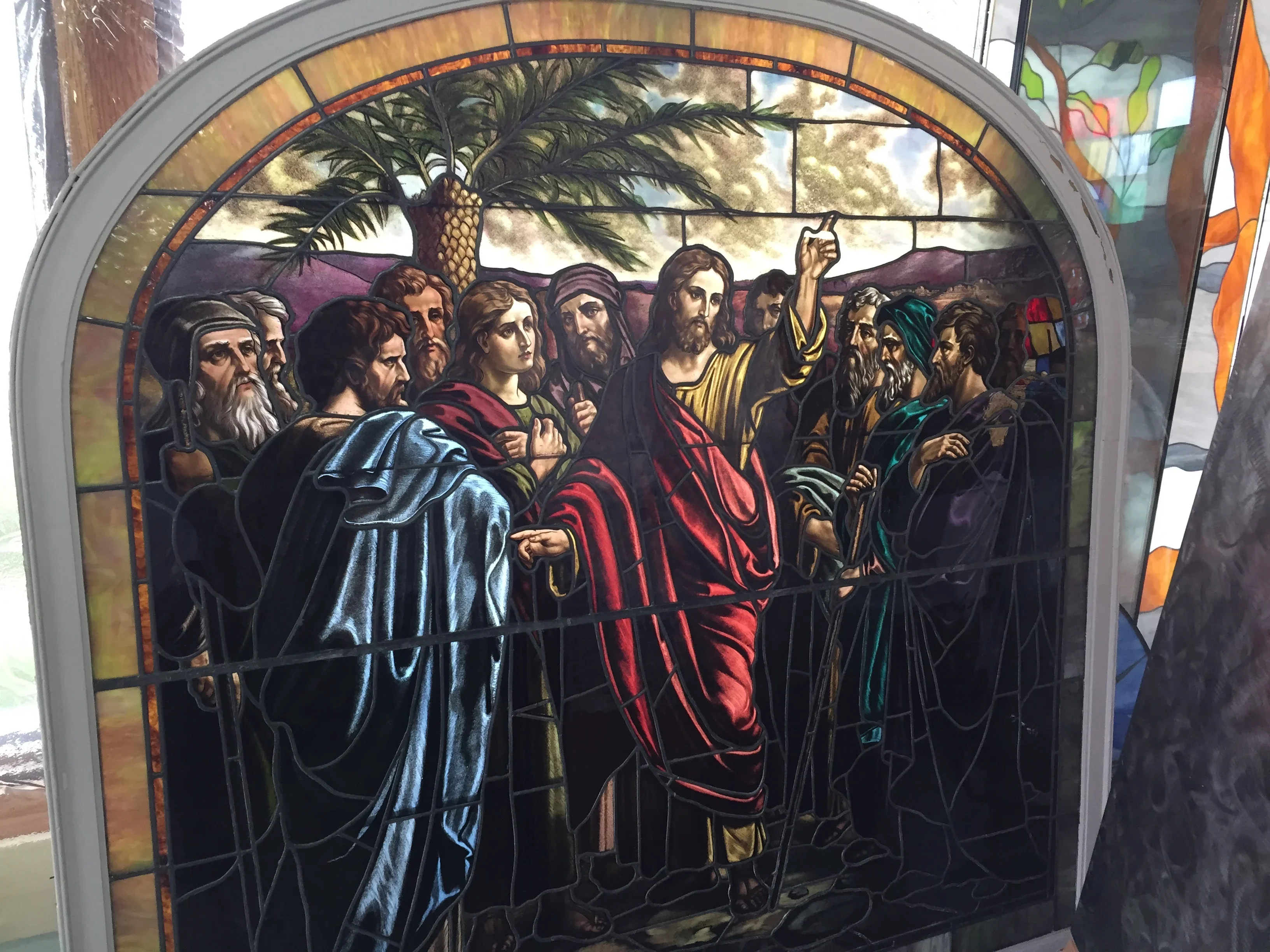 Arched stained glass window depicting religious figures in colorful robes with palm tree and landscape background