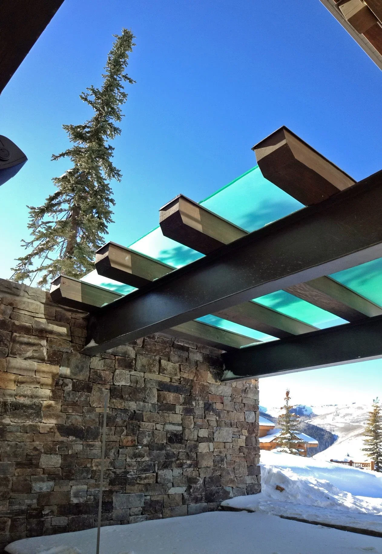 Modern pergola with turquoise glass panels and steel beams over stone wall