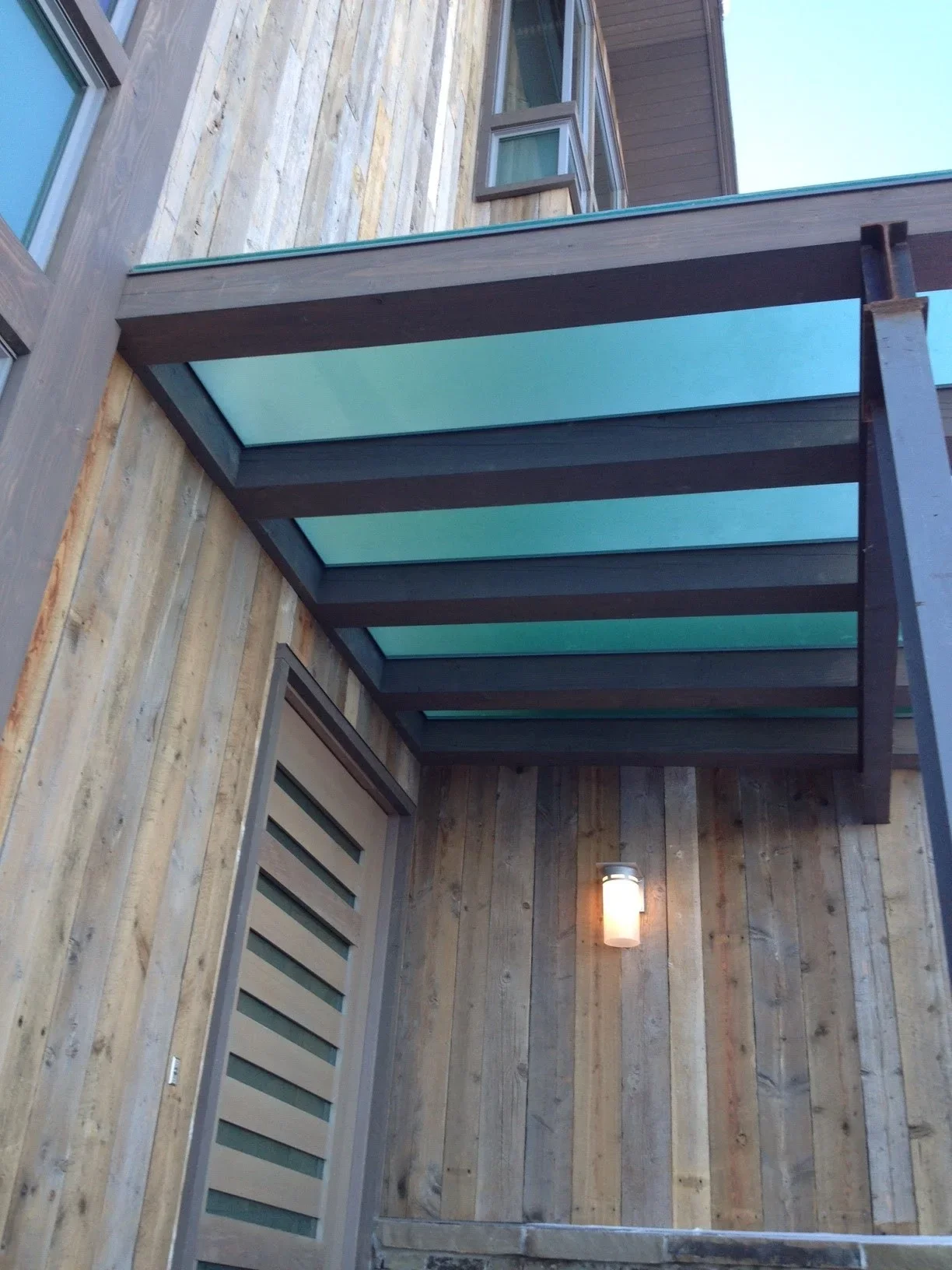 Modern pergola with frosted turquoise glass panels and dark metal frame over wooden entryway
