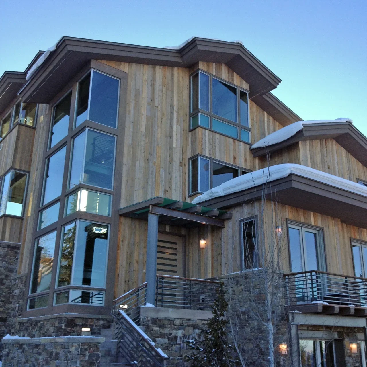 Modern home with large custom windows, wood siding, metal railings, and snow-covered roof